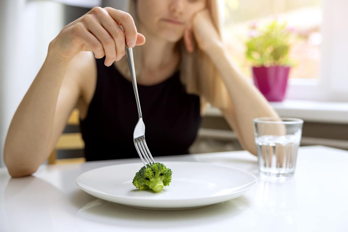 Unhappy young woman holding a fork over a small piece of broccoli on a plate