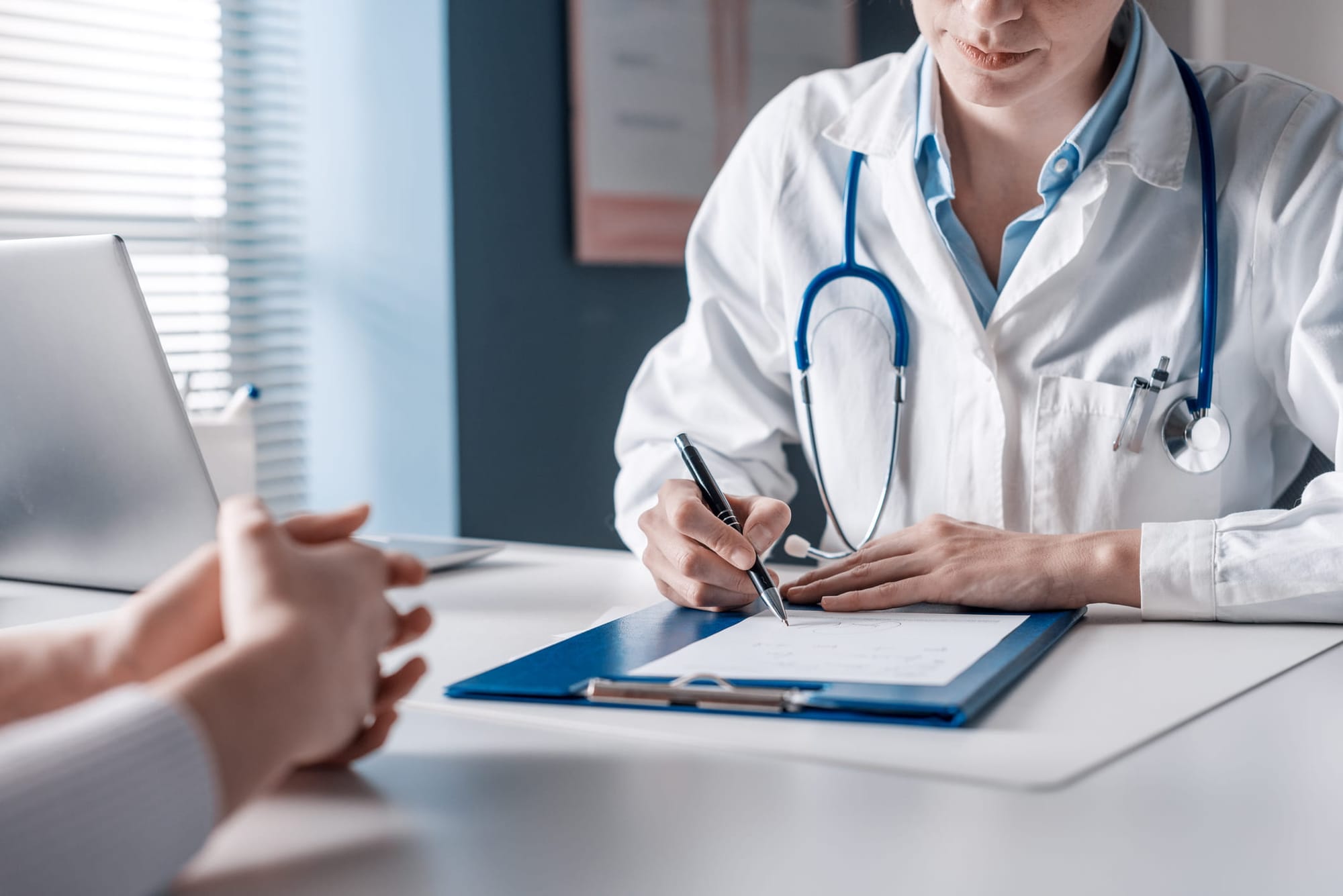 Doctor sitting at desk across from her patient, writing on a clipboard