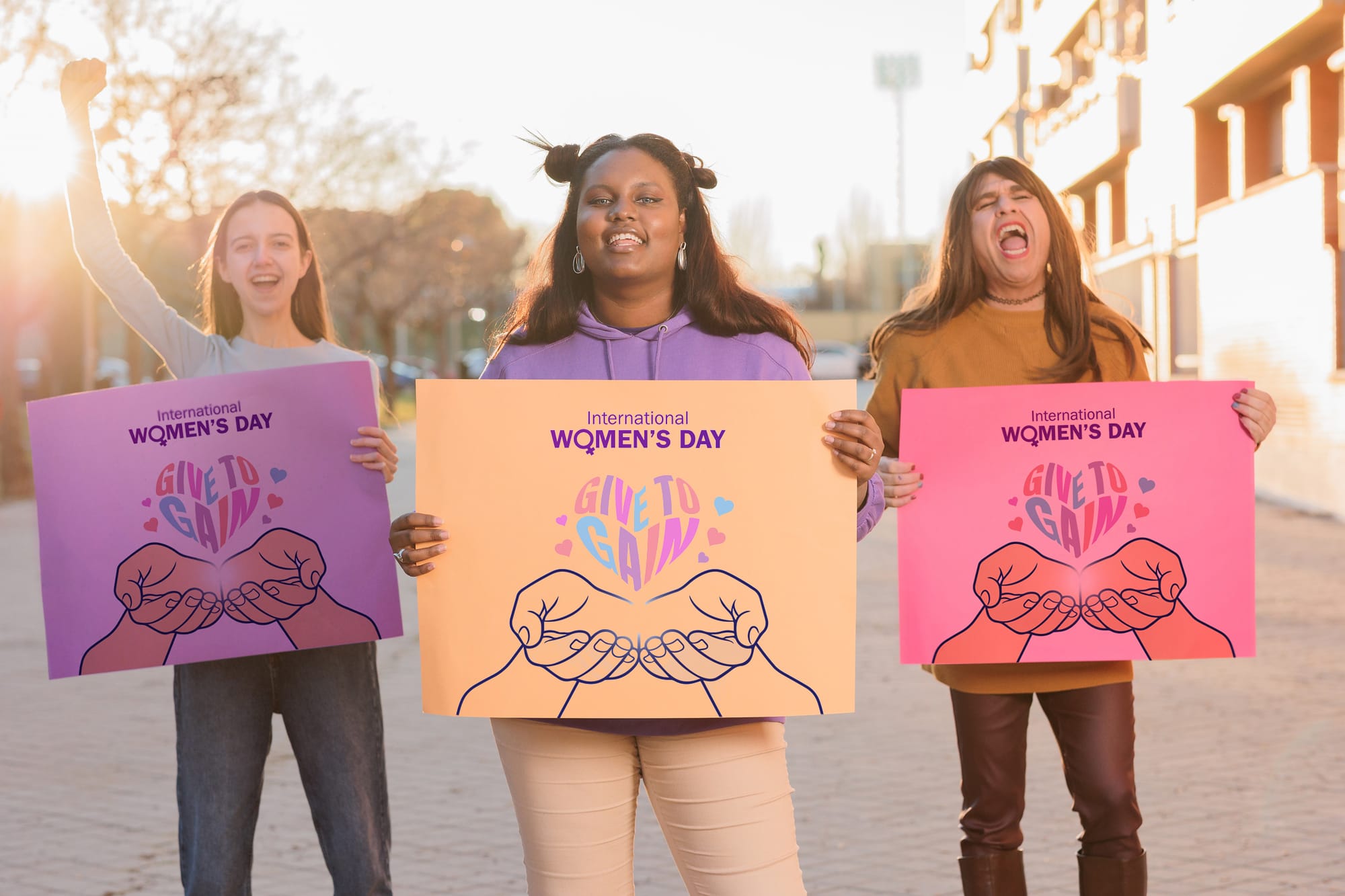 Three smiling trans women holding International Women's Day signs depicting hands cupping a heart shape 