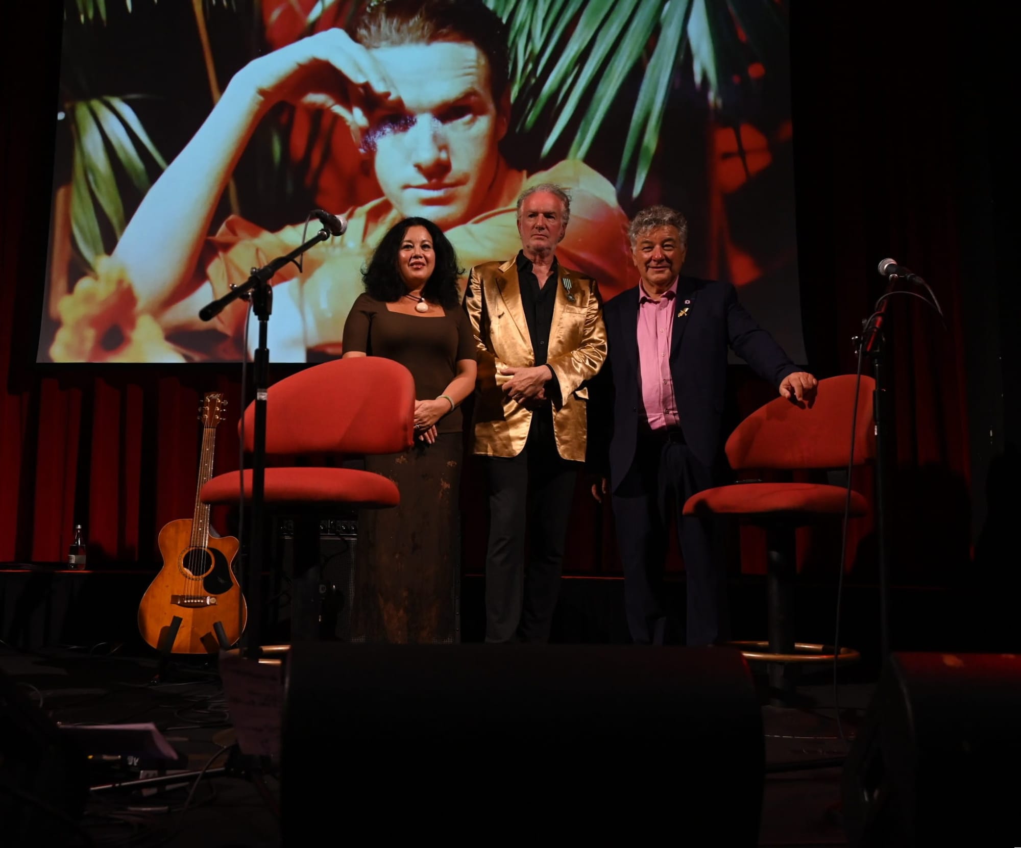 Two men and a woman standing on a stage flanked by chairs and a guitar, with an image of Mick Harvey behind them.