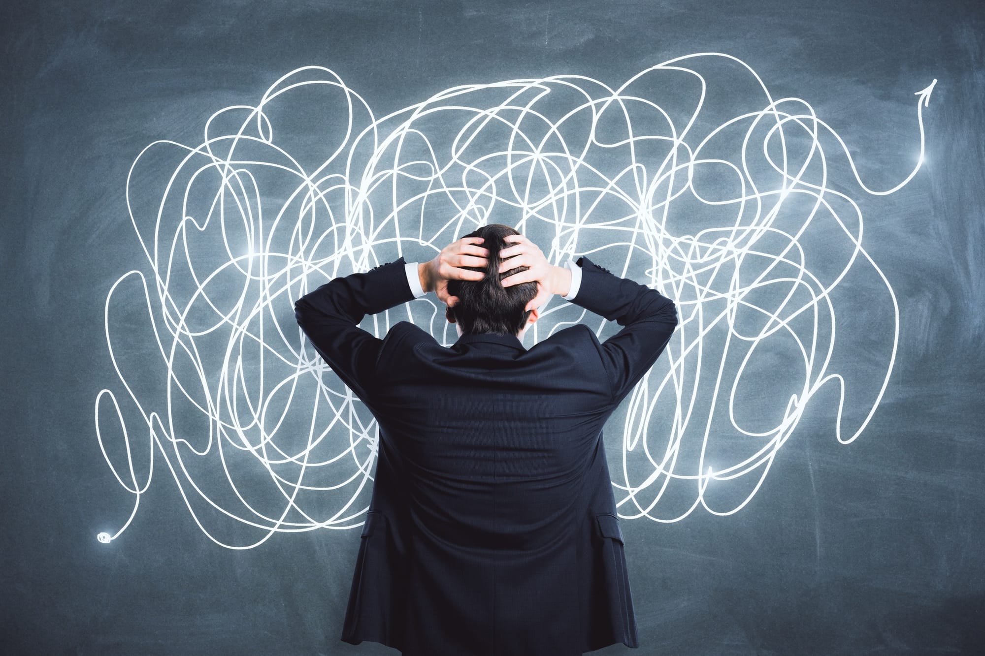 Man facing a blackboard with scribbles across it, with his hands on his head.