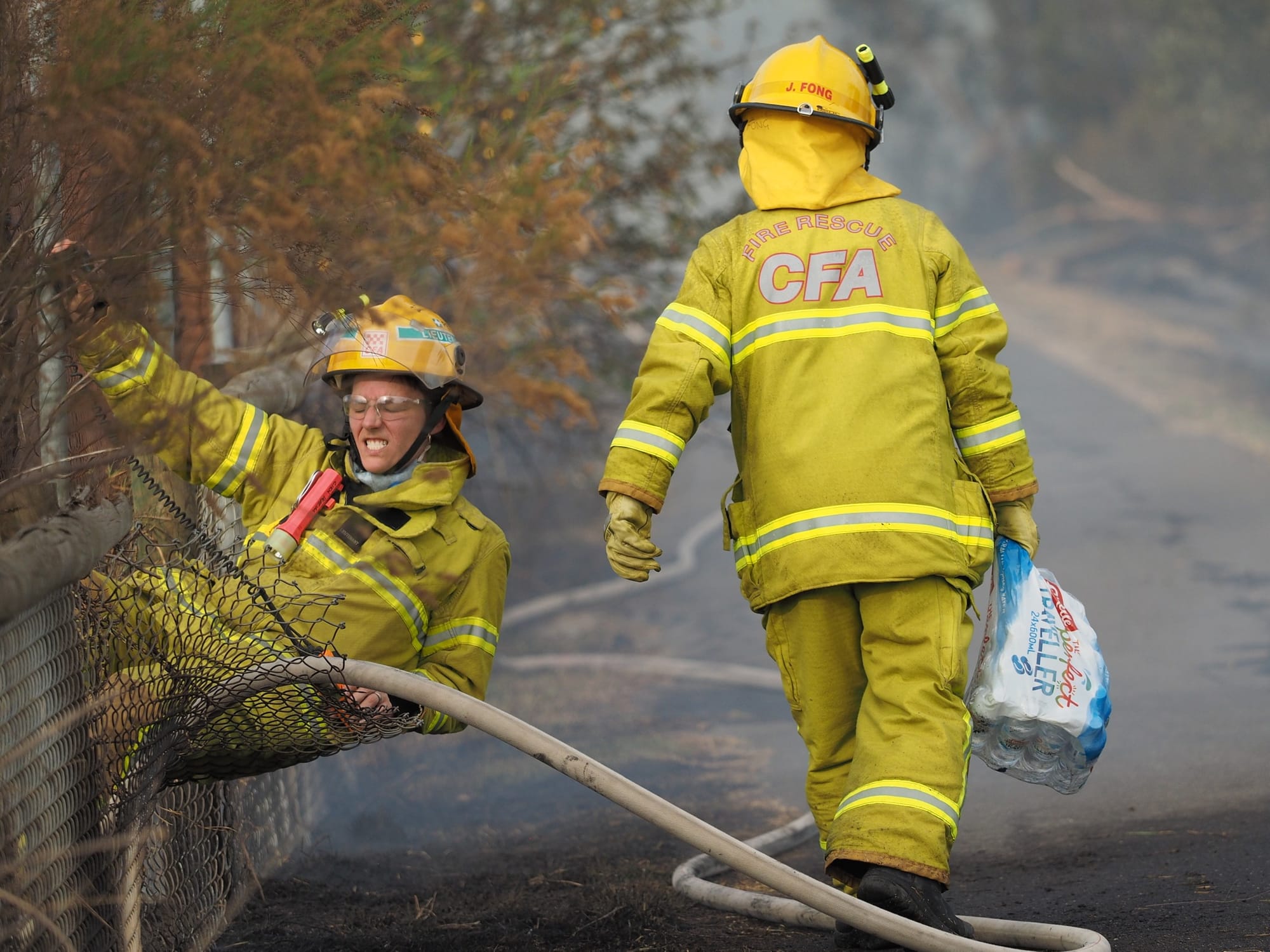 Two firefighters dressed in yellow CFA protective gear at a fence during a bushfire, one straining to climb through a fence