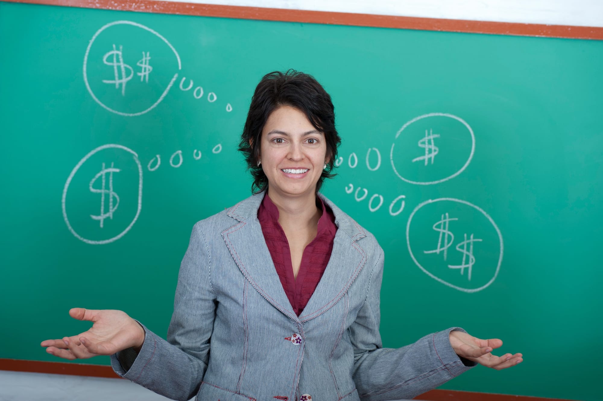 Young female teacher standing in front of blackboard thinking of money.