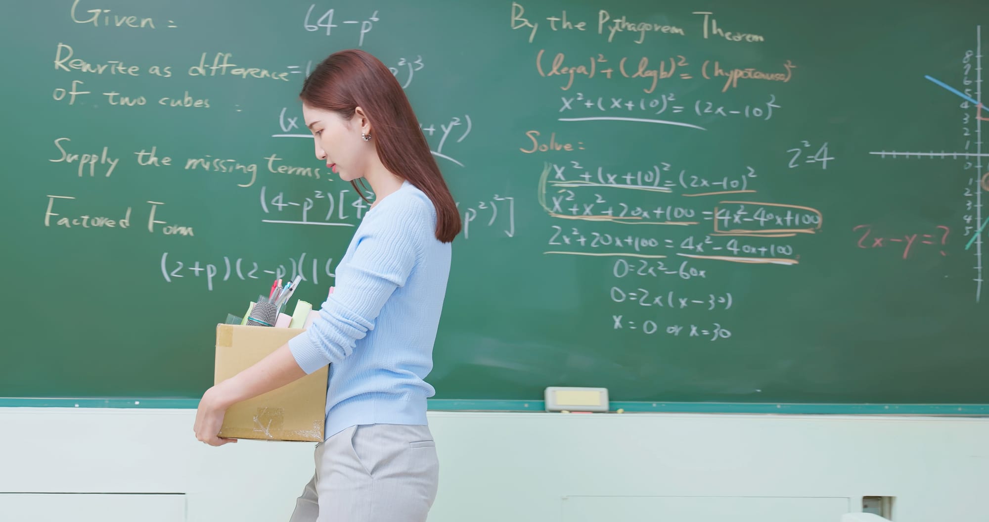 Female teacher with a box of belongings walks passed a blackboard.