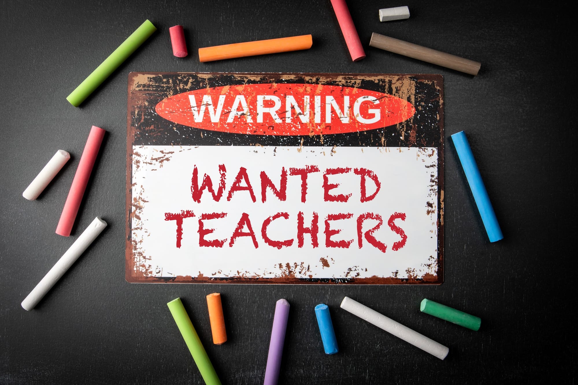 Metal warning sign 'teachers wanted' and coloured pieces of chalk on a dark chalkboard background