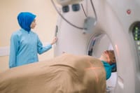 A female Asian nurse in a headscarf preparing a male patient for a scan laying on a CT scanner bed