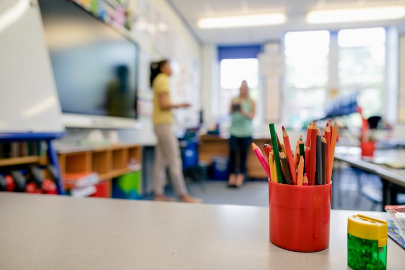 A desk in a classroom with a colourful pot of pencils on the desk next to a pencil sharpener. In the background is two teachers talking on the classroom.