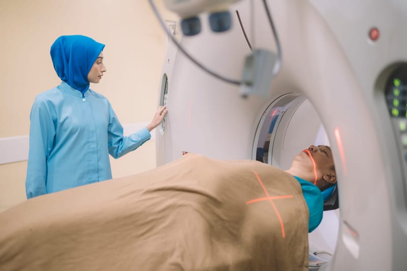 A female Asian nurse in a headscarf preparing a male patient for a scan laying on a CT scanner bed