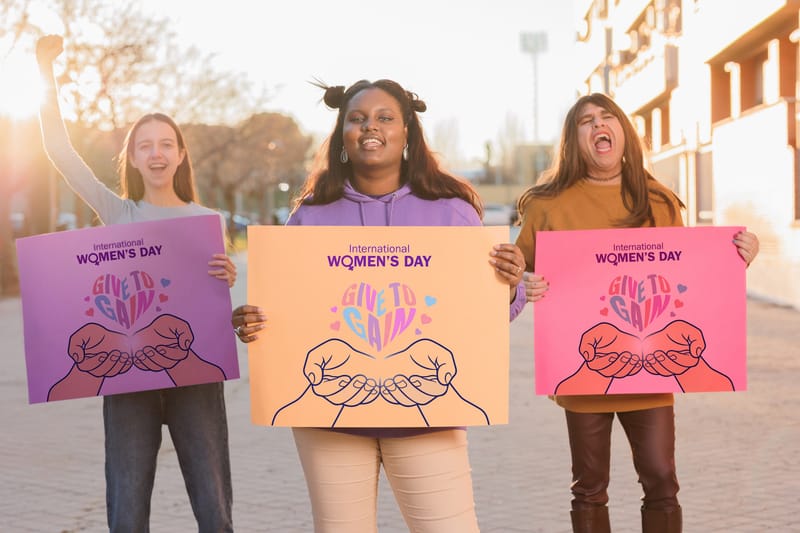 Three smiling trans women holding International Women's Day signs depicting hands cupping a heart shape 