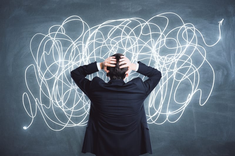 Man facing a blackboard with scribbles across it, with his hands on his head.